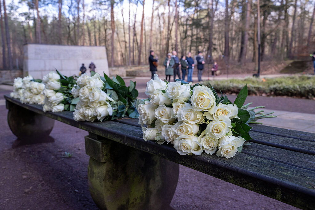 Weiße Rosen liegen auf einer Bank auf dem Heidefriedhof; im Hintergrund sind Menschen zu sehen